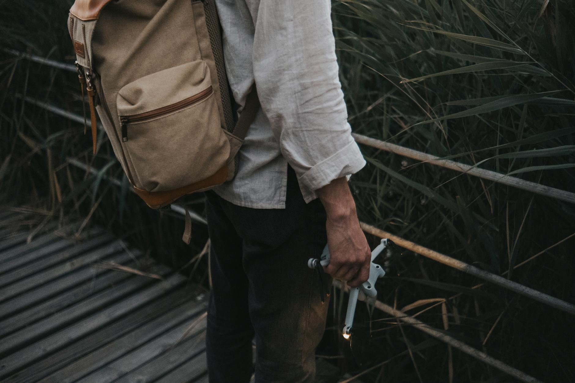 Mężczyzna z plecakiem trzyma drona na drewnianym pomoście wśród traw