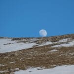 Księżyc wschodzący nad ośnieżonym stokiem górskim na tle błękitnego nieba