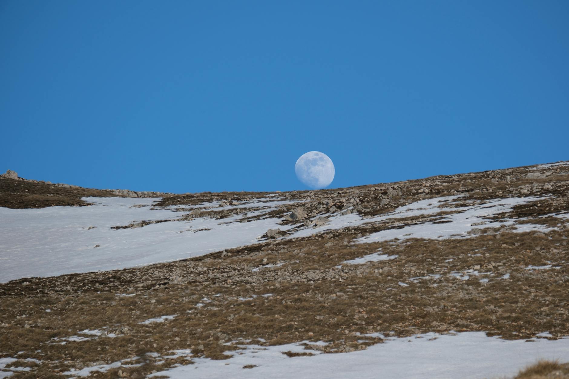 Księżyc wschodzący nad ośnieżonym stokiem górskim na tle błękitnego nieba